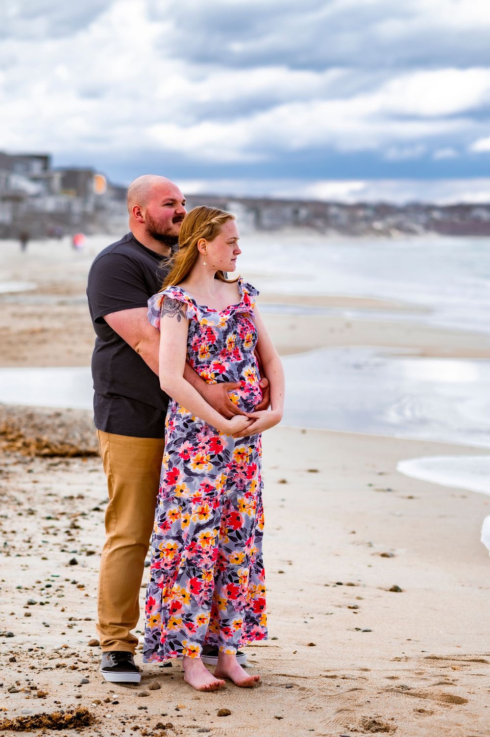 A couple embraces while standing on a beach with cloudy skies and ocean waves.