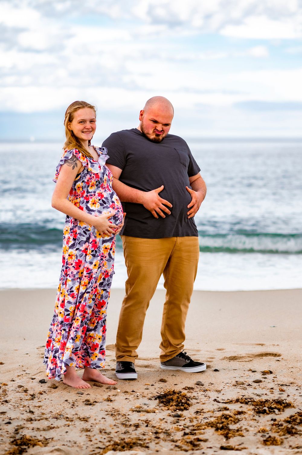 A couple shares a lighthearted moment on a sandy beach with waves behind them.