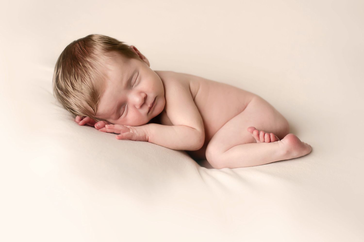 Peaceful sleeping newborn baby curled up on cream colored background.