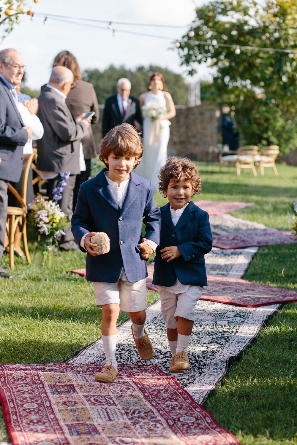 Two young boys in navy blazers walking down Persian rugs at an outdoor wedding ceremony.
