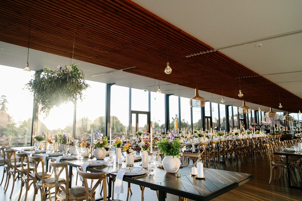 Long banquet tables set for wedding reception in modern venue with wooden ceiling and large windows.