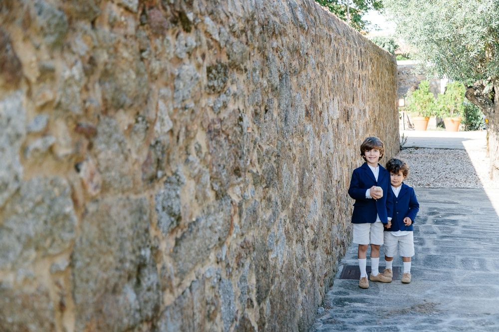 Two young ring bearers in matching outfits stand against a rustic stone wall.