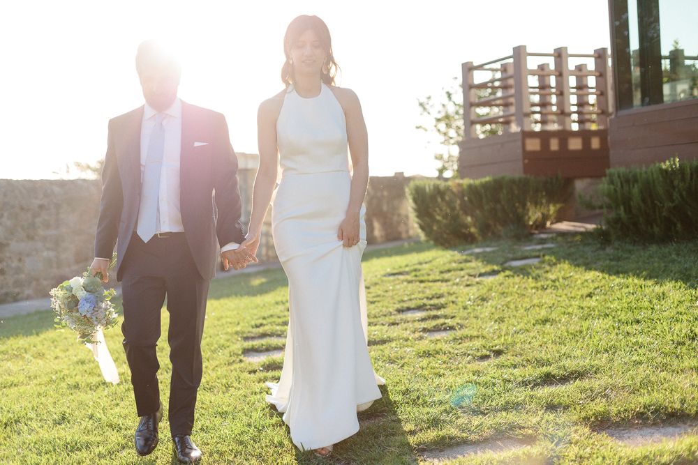 A couple in wedding attire walks hand in hand across a sunlit grassy lawn during golden hour.