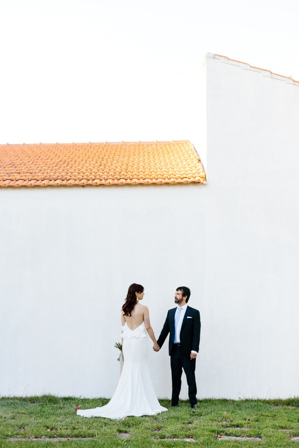 A couple in wedding attire stands intimately against a white wall with terracotta roof tiles.