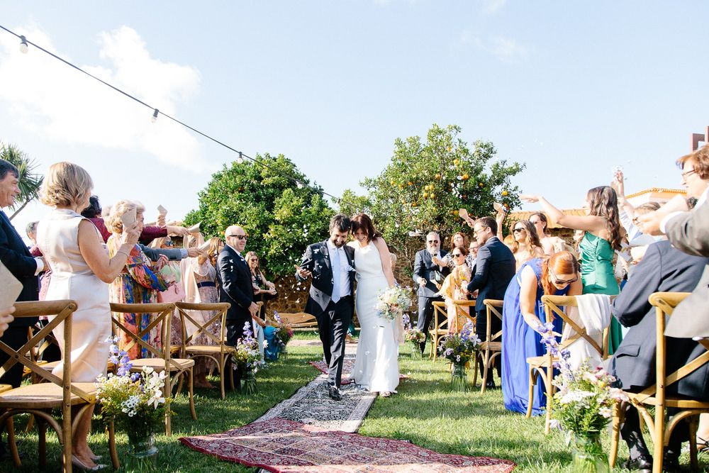 Wedding ceremony outdoors with guests seated on wooden chairs along a boho-style aisle runner.