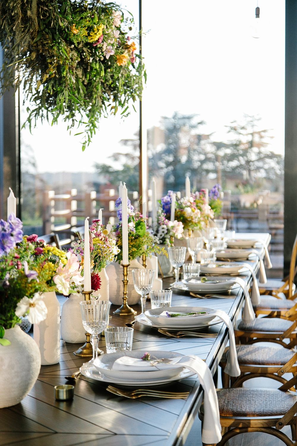 Elegant table setting with hanging floral arrangements, candles, and fine china against window view.