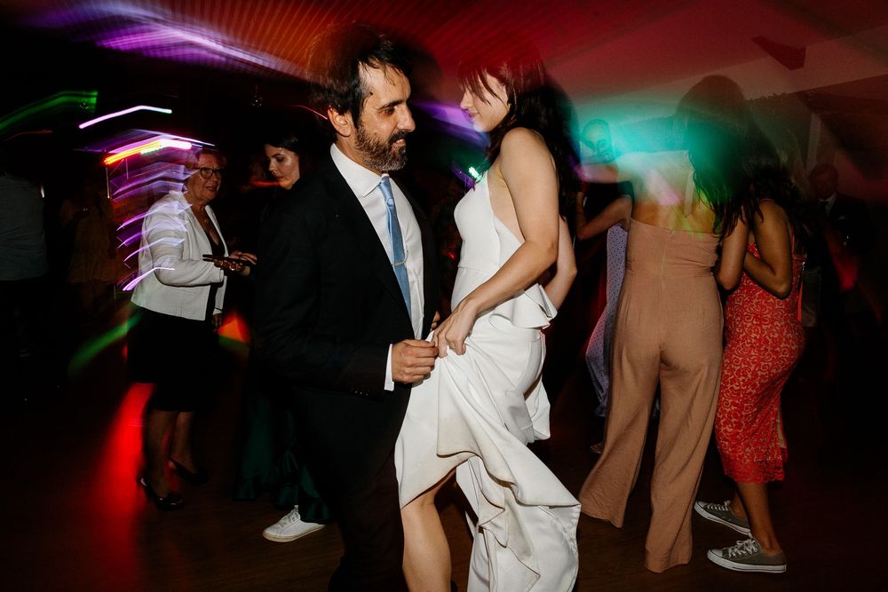 A couple dances together at a wedding reception with colorful lighting and guests in the background.