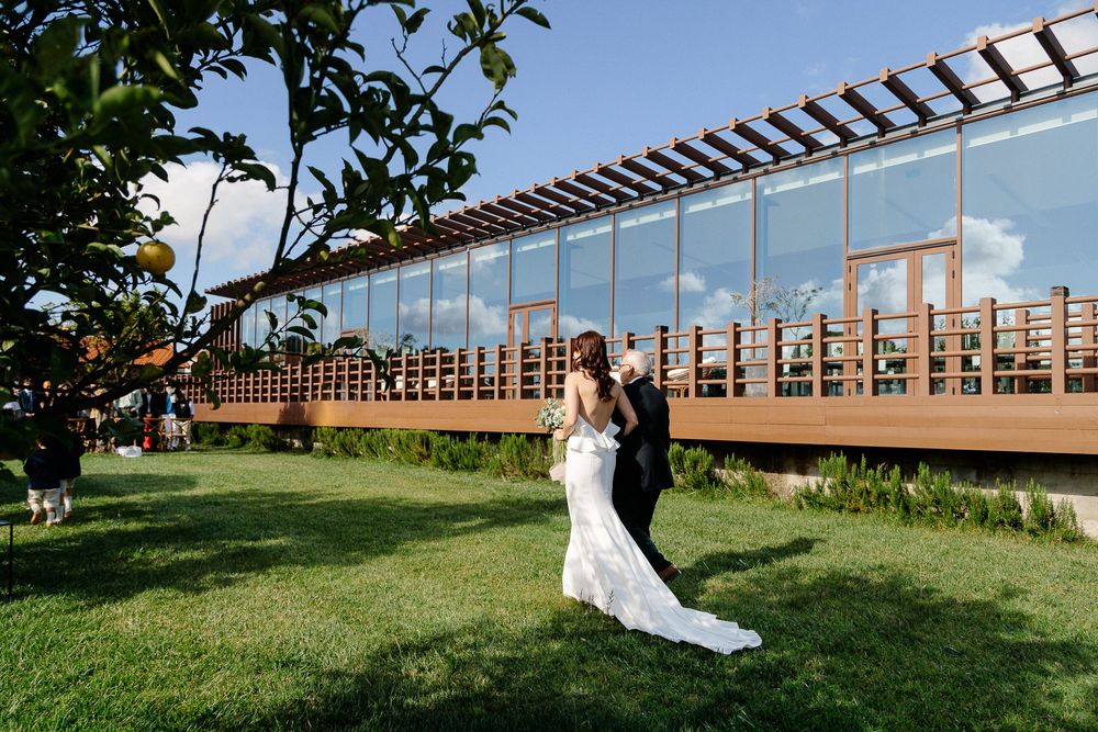 A wedding dress flows in the breeze against a modern wooden pergola structure and green lawn.