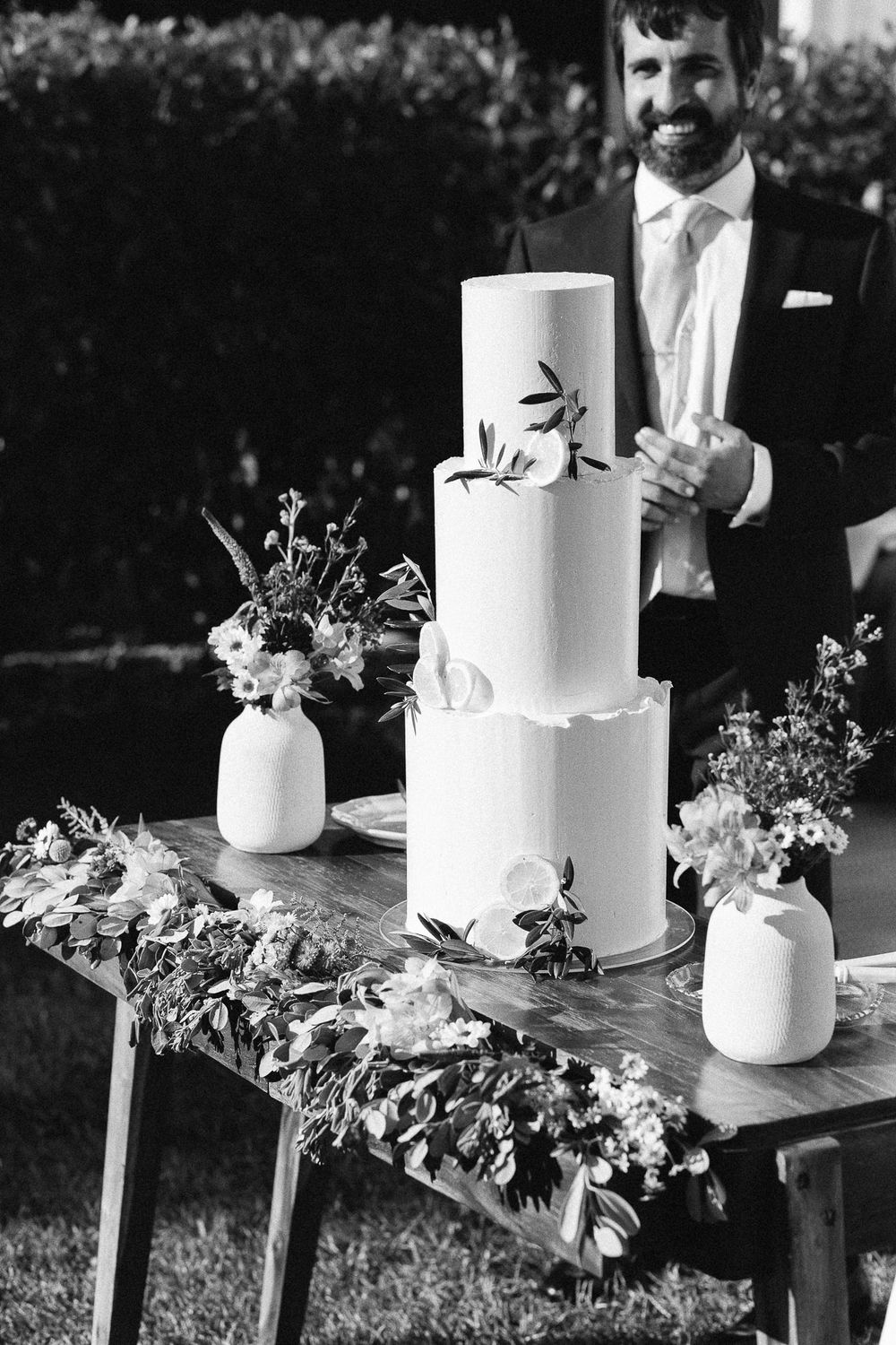 A minimalist white tiered wedding cake decorated with delicate greenery and small white floral arrangements.