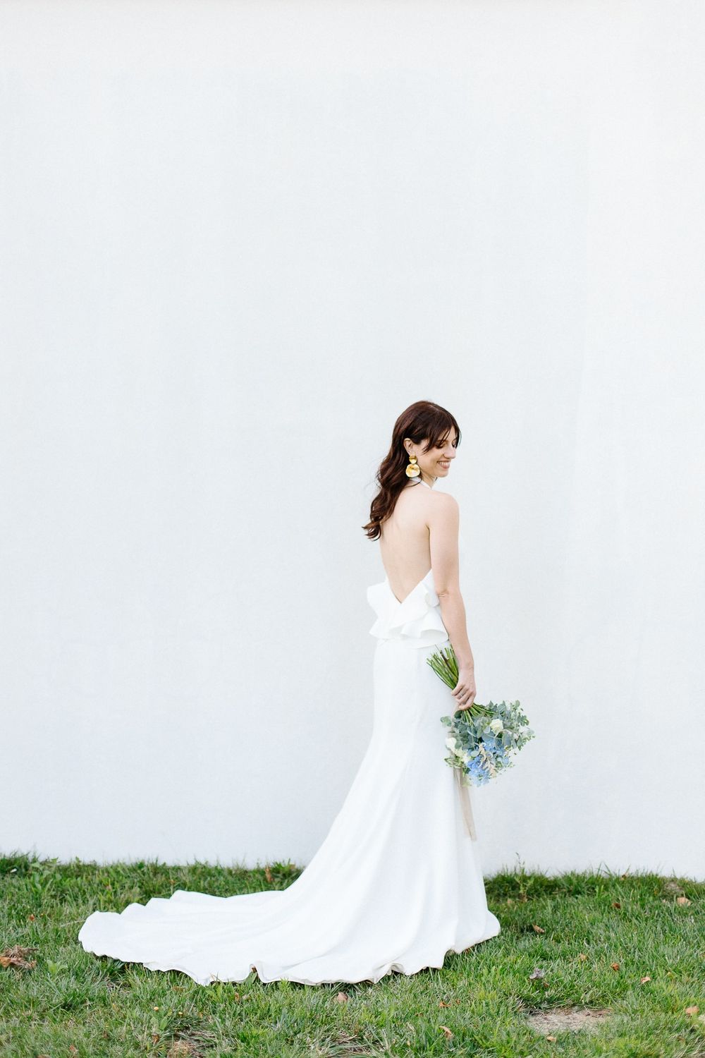 A bride in a sleek white gown poses with bouquet against a minimalist white wall.