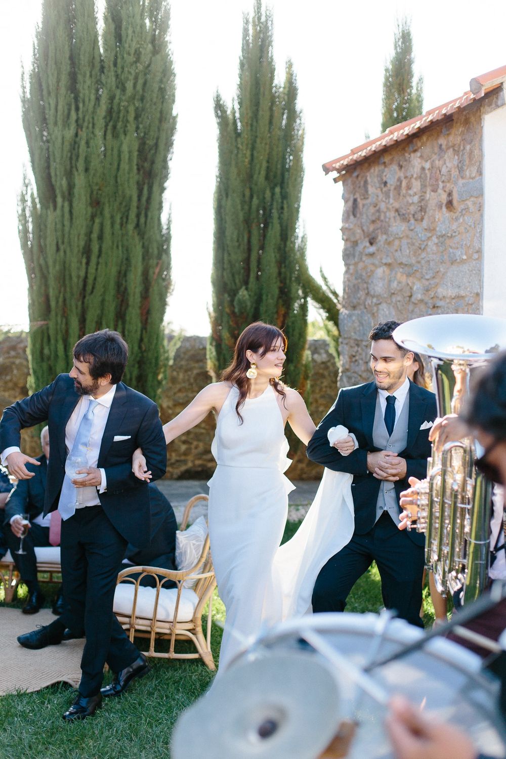 Wedding celebration with dancers and musicians performing in a garden setting with tall cypress trees.