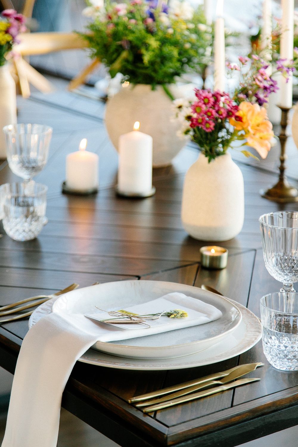 Elegant table setting with white candles, colorful flower arrangements, and crystal glassware on dark wood.