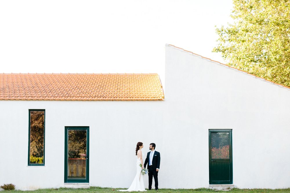 A bride and groom pose in front of a white building with green doors and terracotta roof.