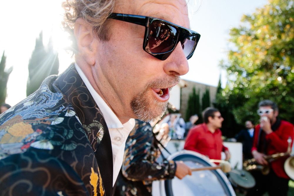 Musician in sunglasses and patterned jacket performing energetically at an outdoor event.
