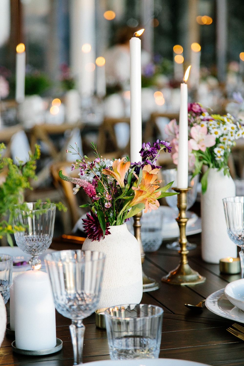 Romantic table setting with tall white candles, colorful wildflowers, and crystal glasses on dark wood surface.