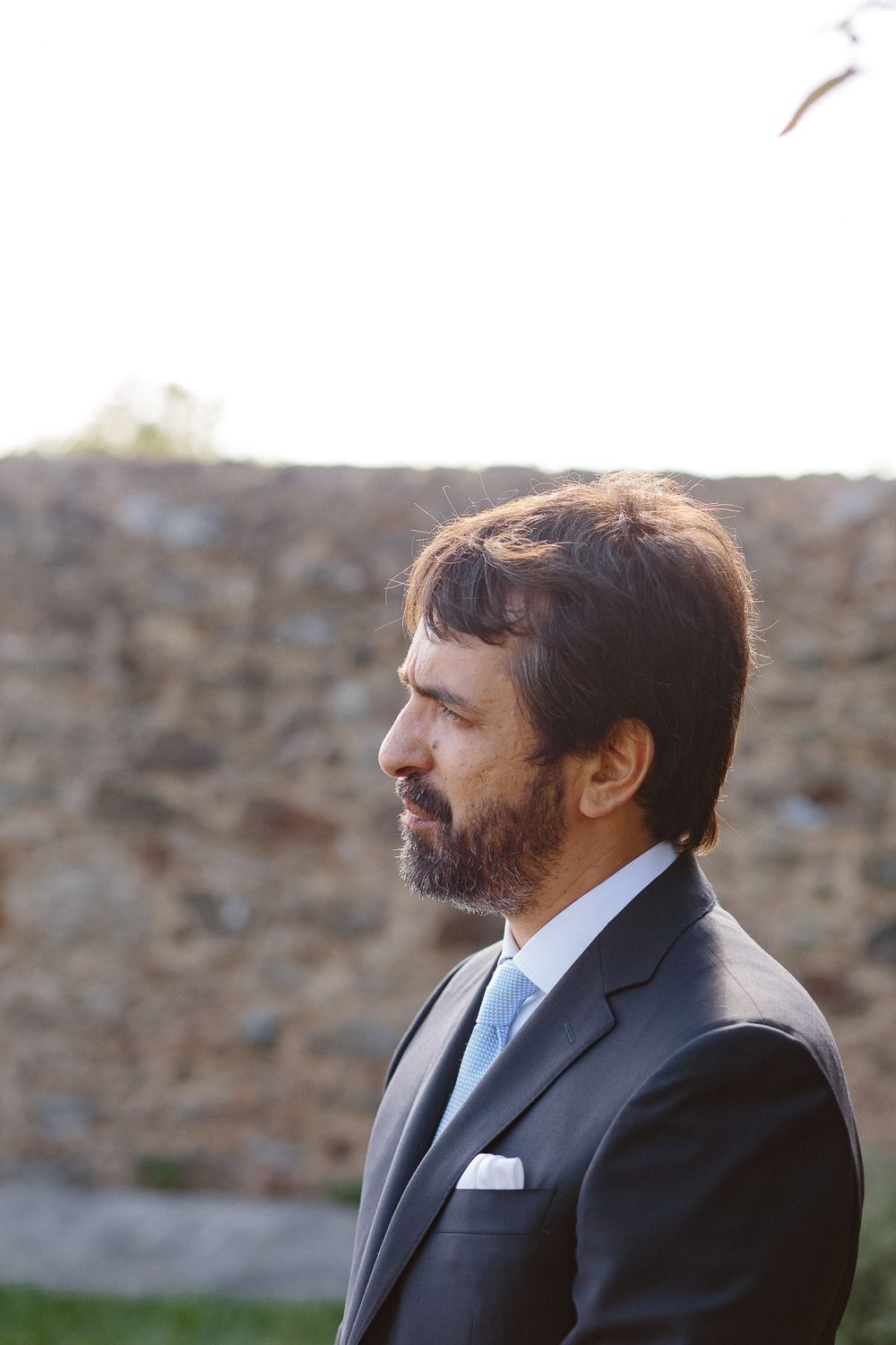 Profile portrait of groom in navy suit against stone wall at sunset.