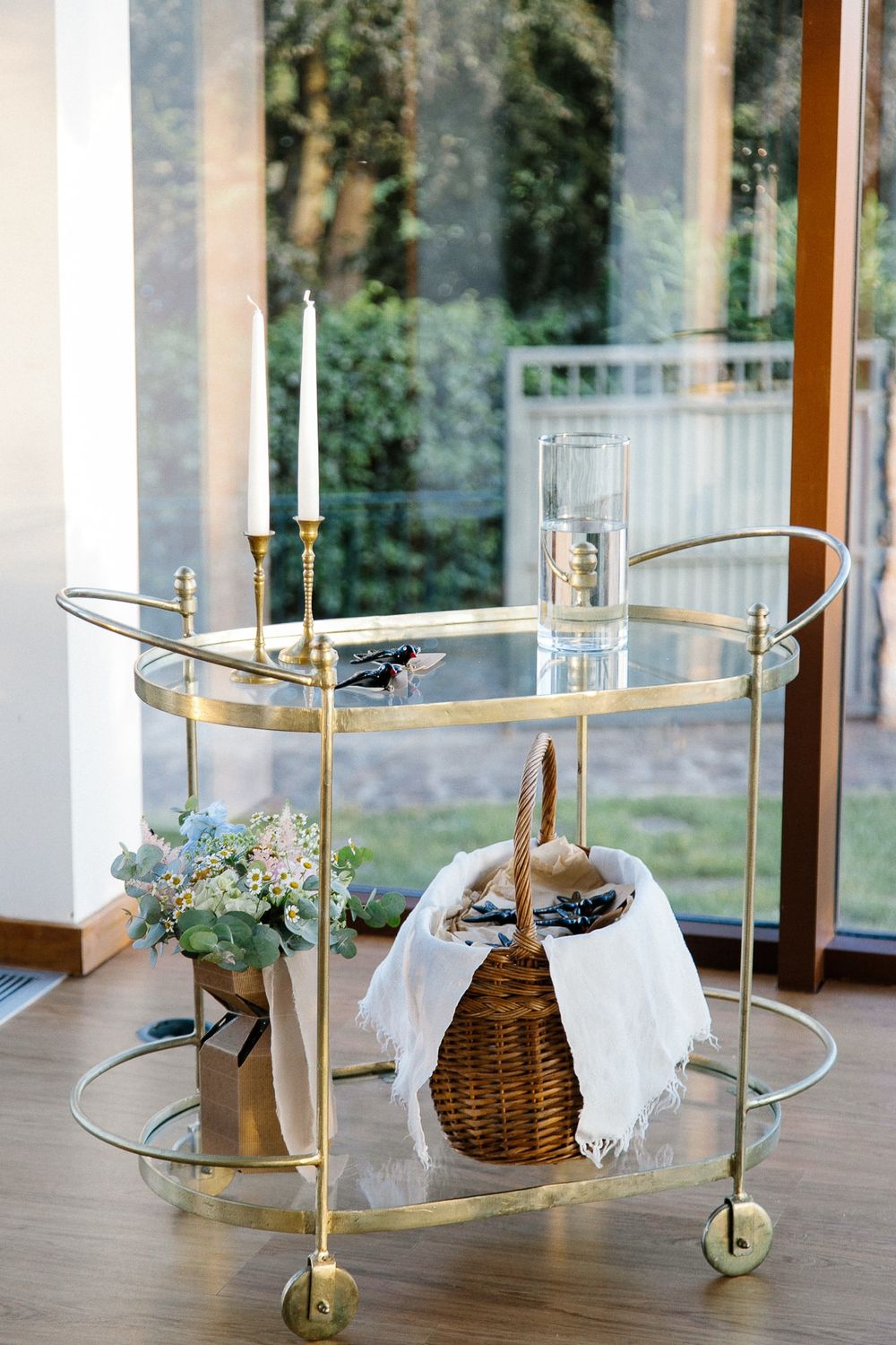 Elegant gold bar cart with glass shelves displaying candles, flowers and a woven basket against a window backdrop.