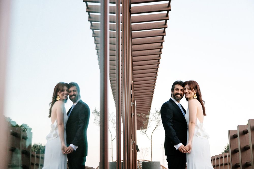 Creative mirror reflection shot of couples in formal attire against modern architectural backdrop.