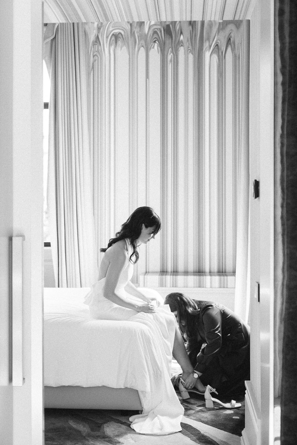 A black and white photo shows final wedding dress preparations in a light-filled room.