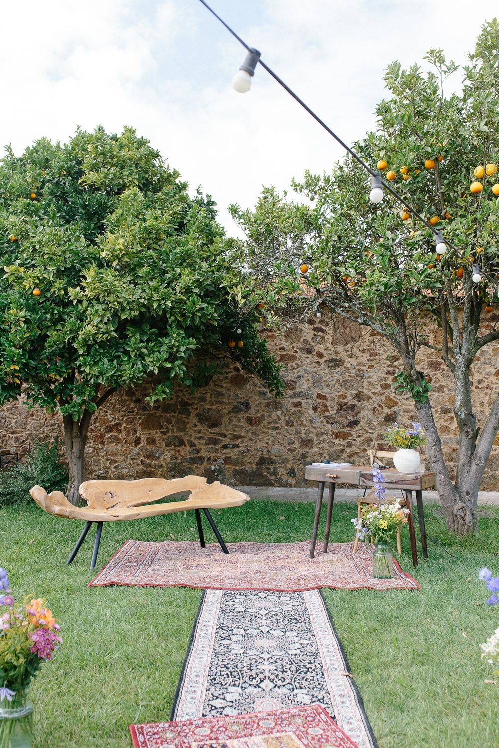 Garden lounge area with string lights, rugs, and citrus trees against stone wall.