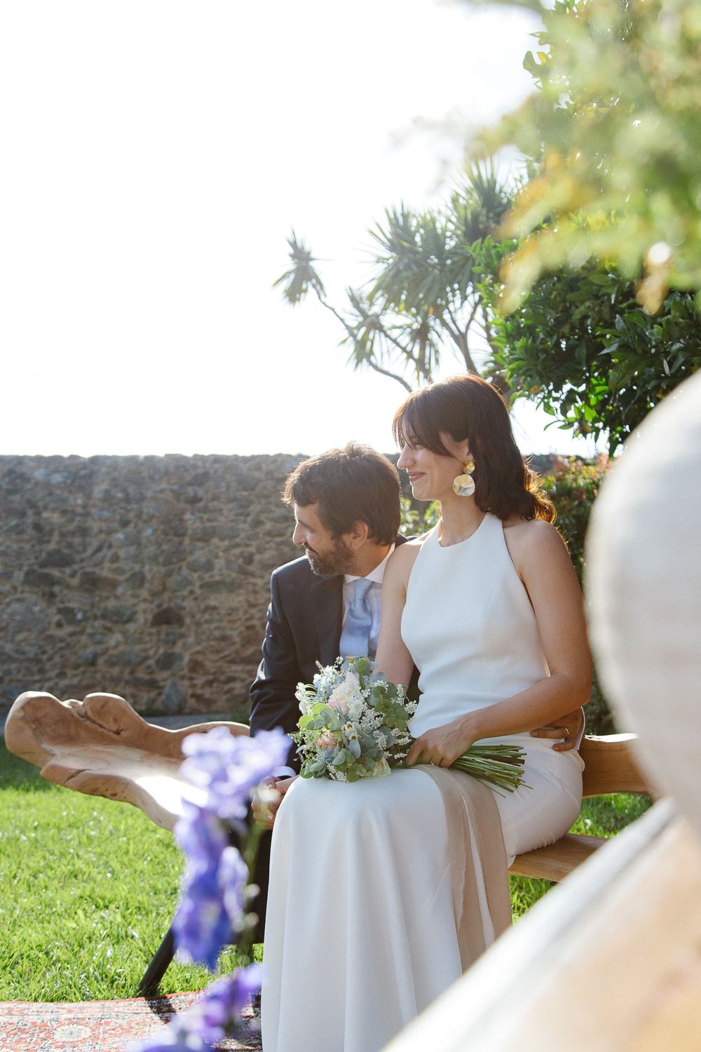 Newlyweds share an intimate moment on a garden bench with white florals and stone wall backdrop.