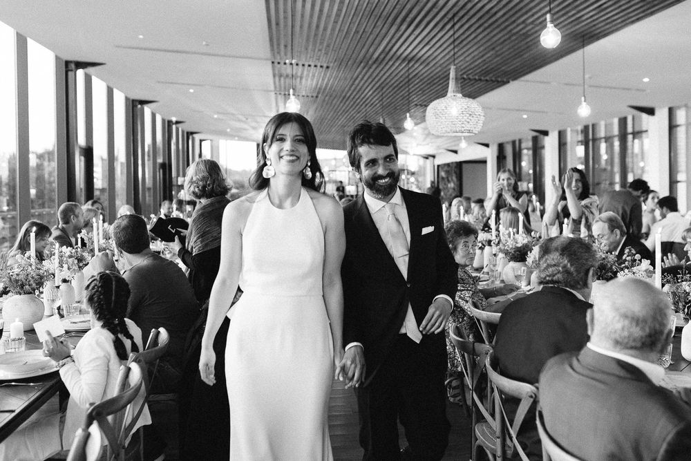 Black and white photograph of reception guests dining in elegant venue with floor-to-ceiling windows.
