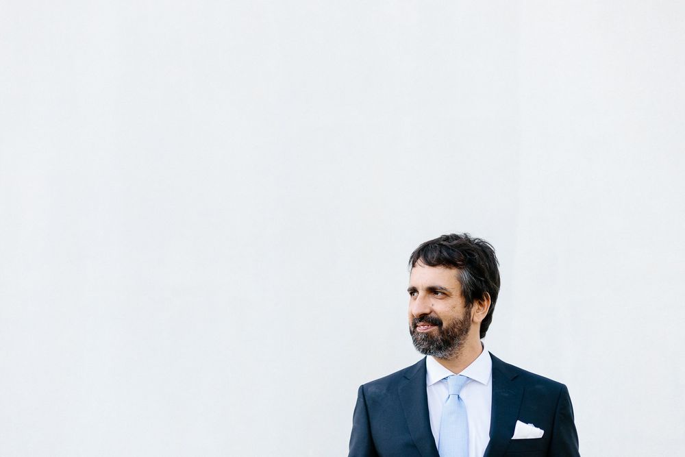 A groom in navy suit and light blue tie poses against a stark white wall.