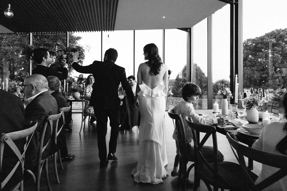 Black and white candid photograph of wedding celebration in modern venue with panoramic windows.