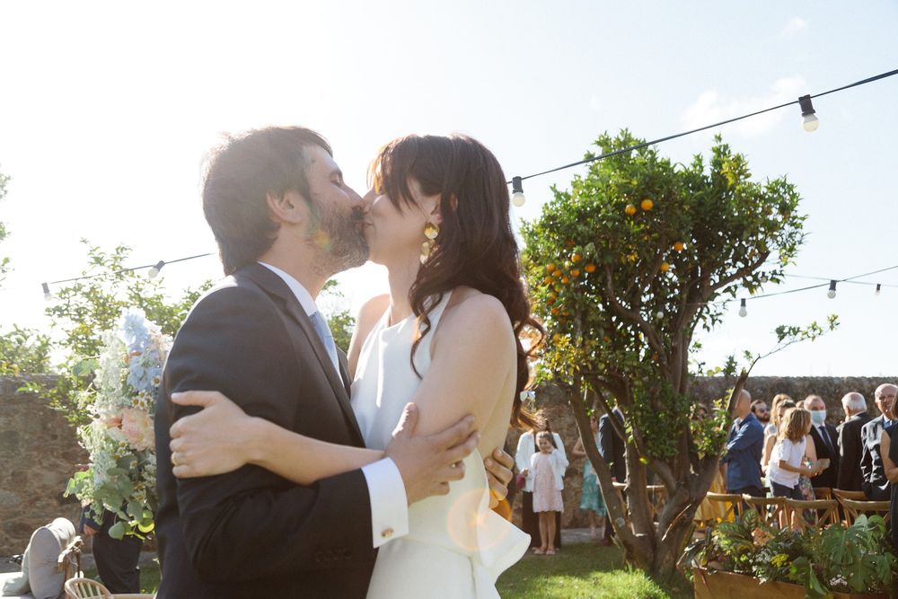 A romantic wedding kiss outdoors during a garden ceremony with string lights and guests in the background.