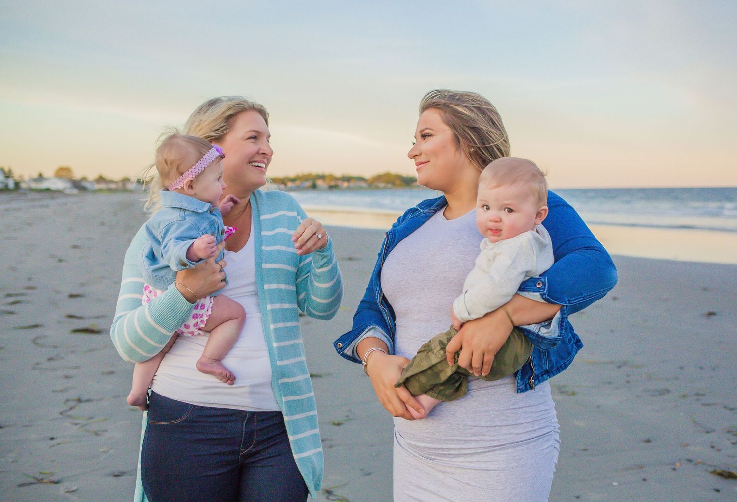 Two mothers holding their babies while sharing a conversation on sunny beach.