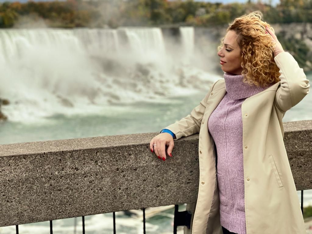 Person in beige coat and purple sweater stands near Niagara Falls on an overcast day.