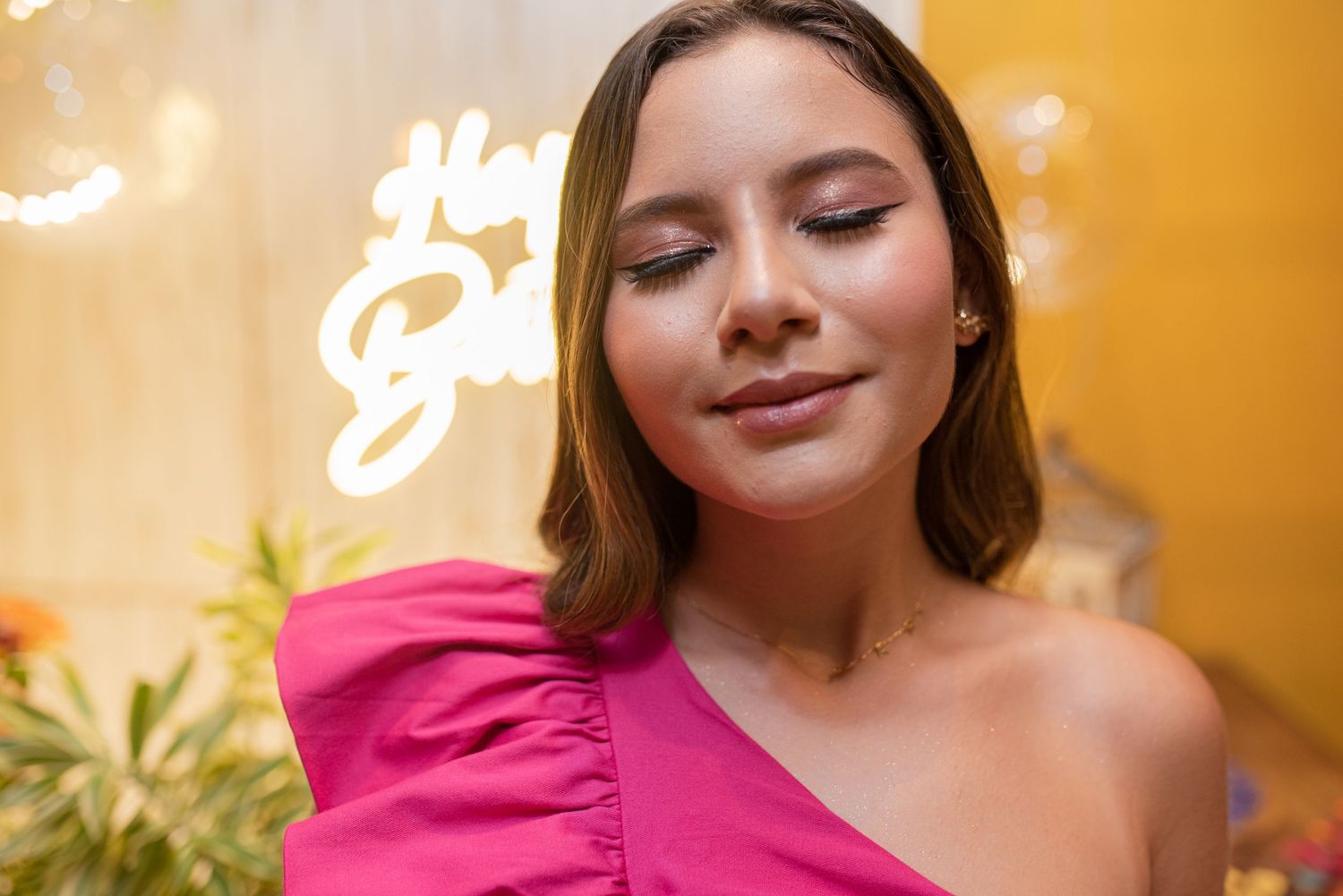 A person in a bright pink one-shoulder top poses against a warm orange background with decorative lighting.