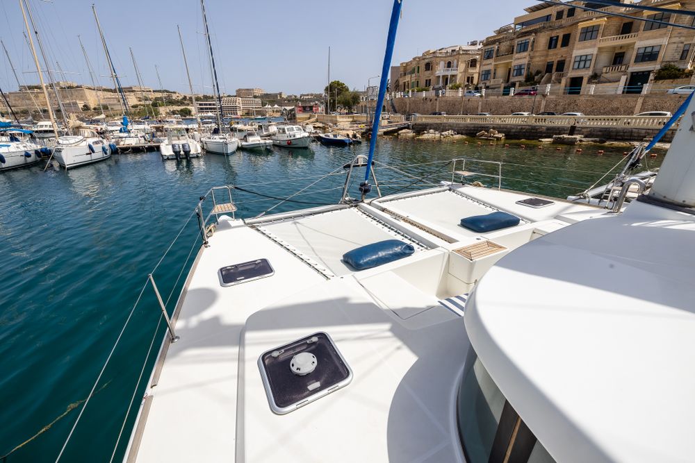 White catamaran deck with blue cushions docked in sunny Mediterranean marina with historic buildings.