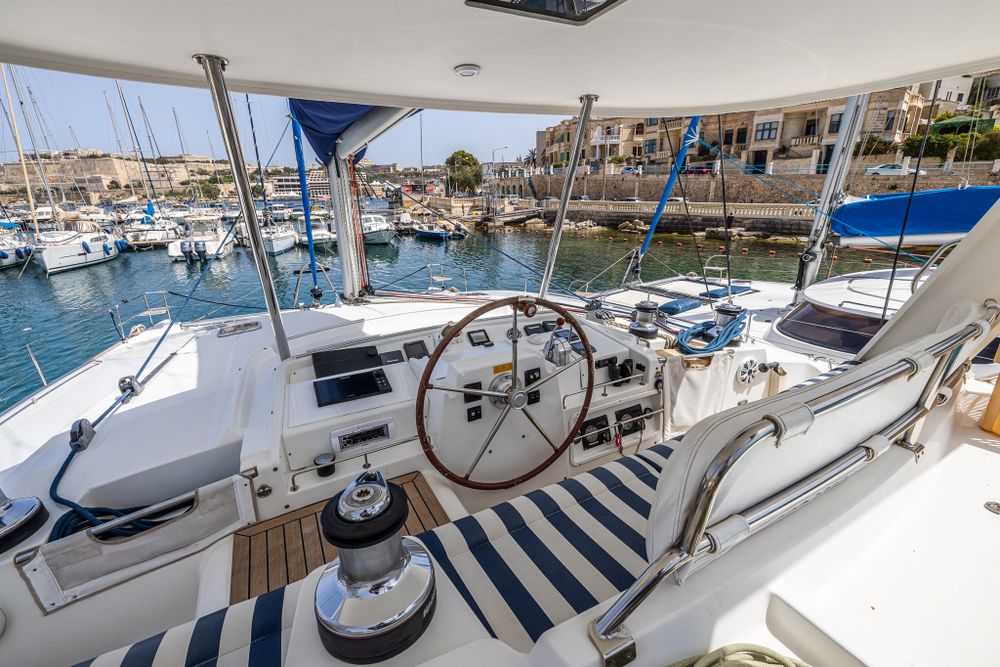 Catamaran helm station with steering wheel, blue striped cushions, and marina view on sunny day.