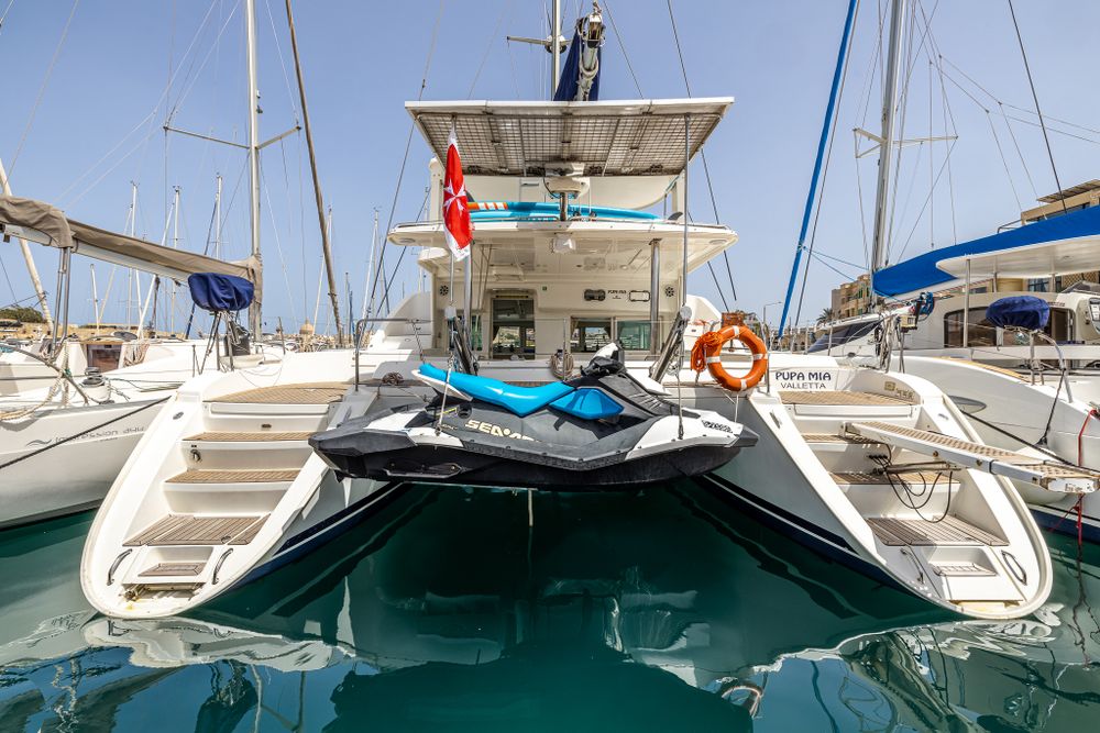 Catamaran stern with Sea-Doo jet ski attached, docked in calm blue marina on sunny day.
