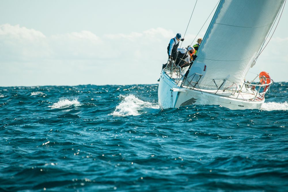 Sailboat heeling sharply in rough blue ocean waves with crew members working the sails.