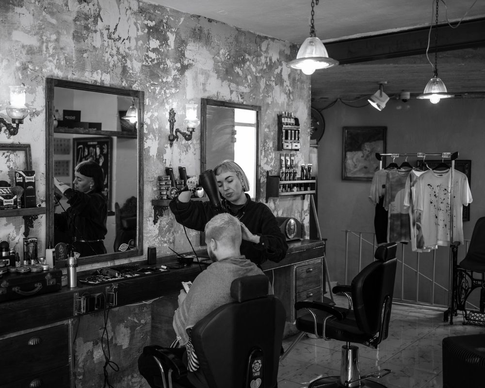 Black and white photo of barber styling client hair in vintage rustic barbershop with mirrors and pendant lights.