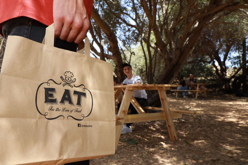 Person holding brown paper EAT branded takeaway bag outdoors with trees and picnic table in background.
