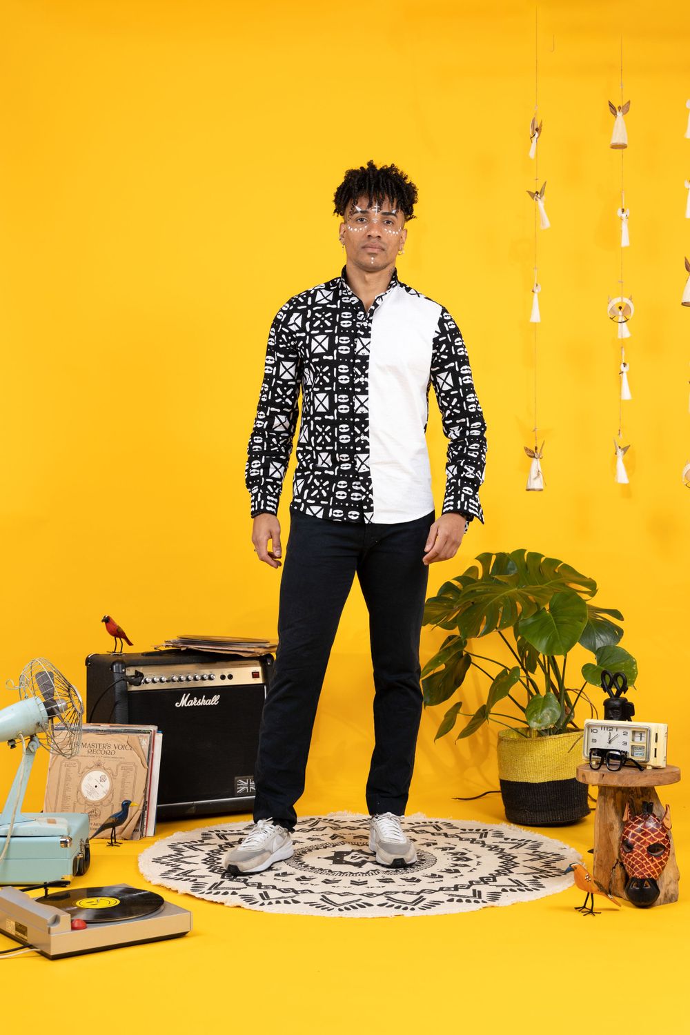 A stylish man poses in a black and white patterned shirt with black jeans against a vibrant yellow background with retro decor.