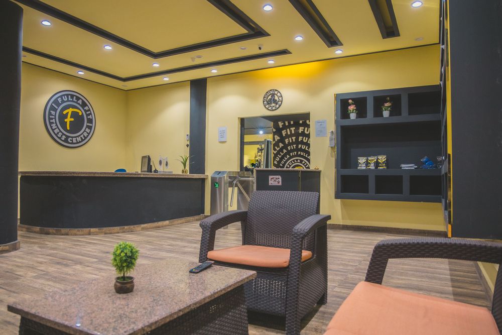 Modern gym reception area with yellow walls, dark wicker chairs, marble table, and a circular logo on the wall.