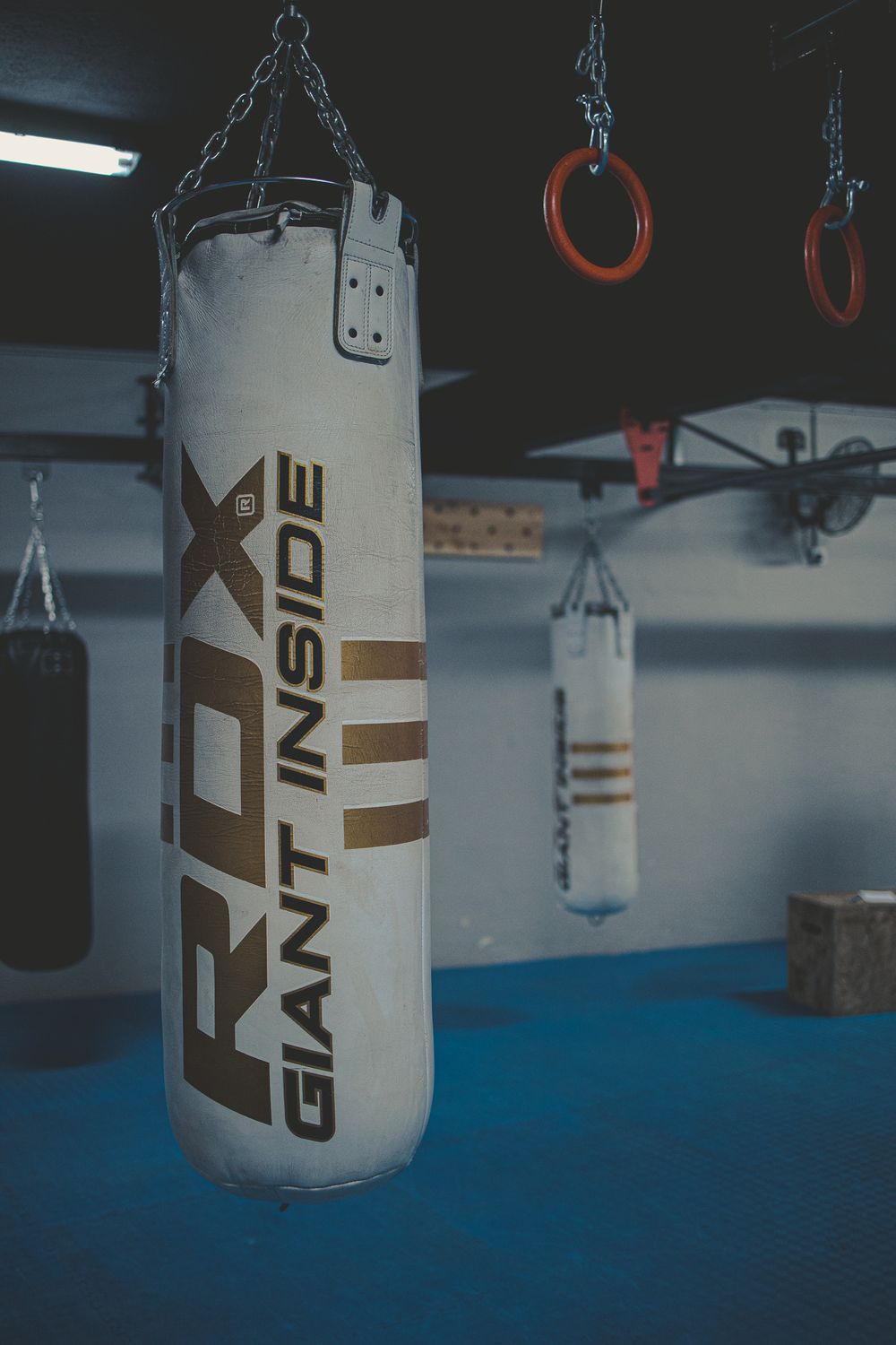 RDX Giant Inside white heavy punching bag hanging in dark boxing gym with gymnastic rings overhead.