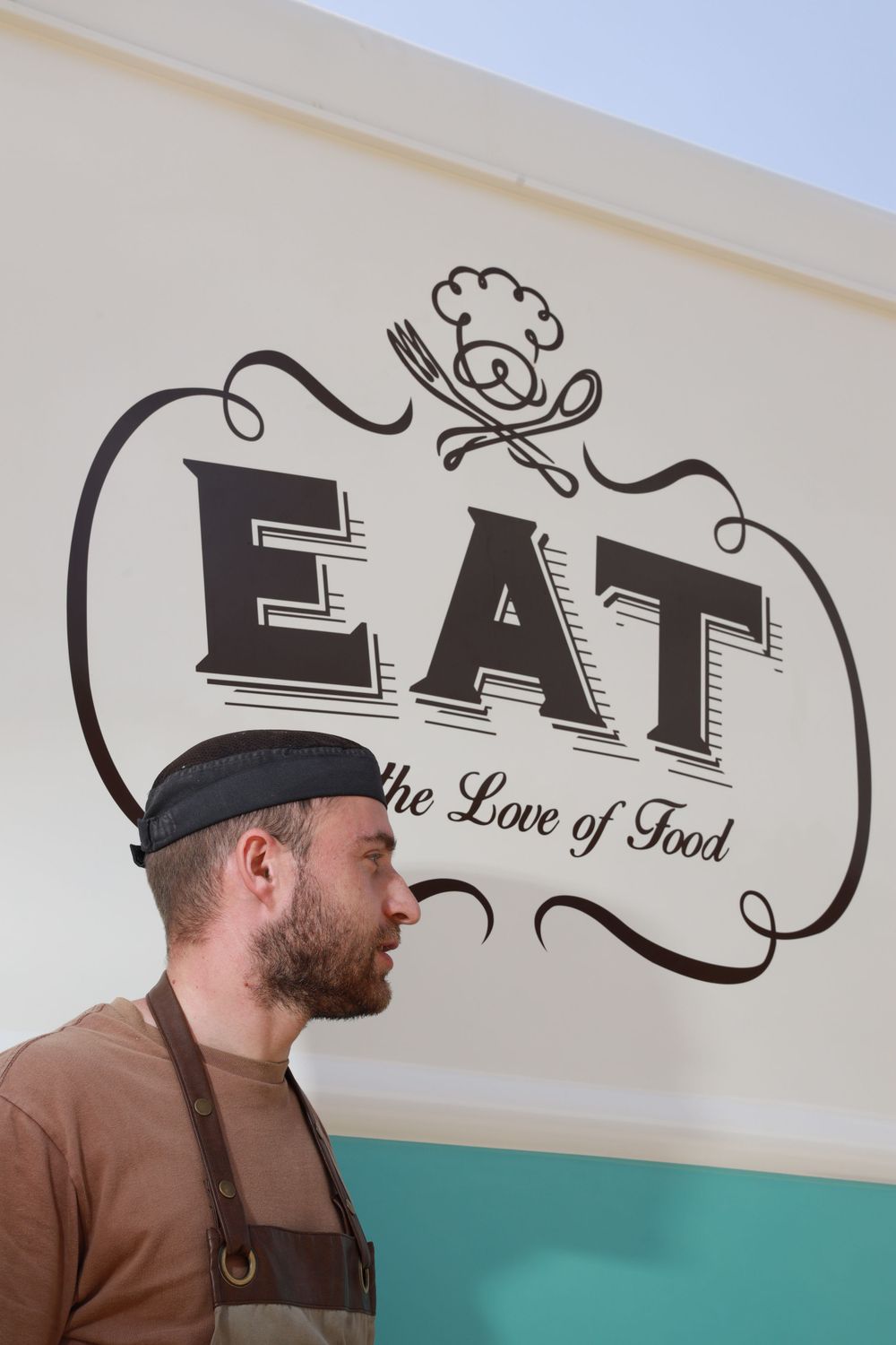 Bearded male chef in black cap and apron standing beside EAT the Love of Food food truck signage.