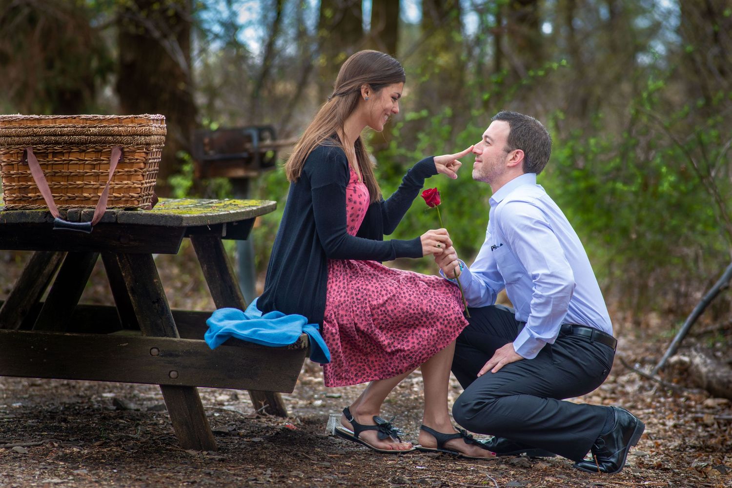 A romantic proposal scene outdoors at a rustic picnic table in a wooded setting with a wicker basket nearby.