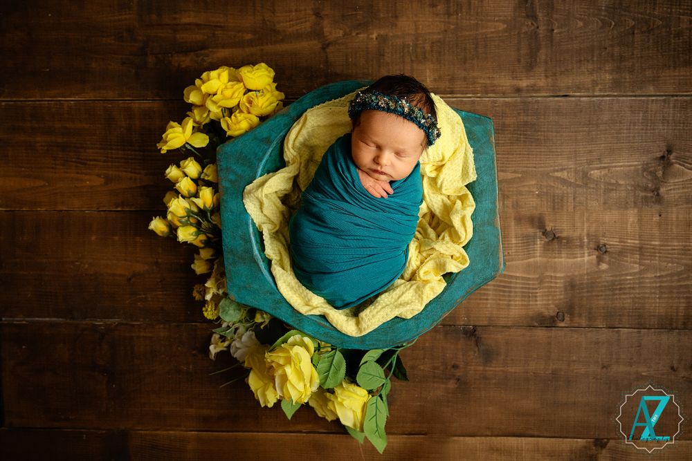 Newborn wrapped in teal swaddle surrounded by yellow flowers in wooden bowl setting.