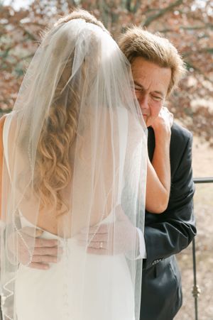 Emotional family member wipes tears while embracing the bride in a white gown and veil outdoors.