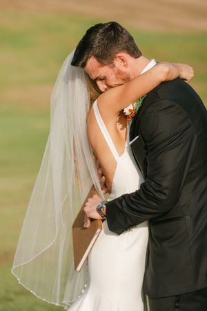 Emotional groom embracing bride from behind during wedding first look, both wearing formal attire outdoors.