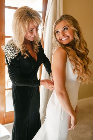 Smiling bride in white gown gets help from her mother in a black velvet dress on wedding morning.