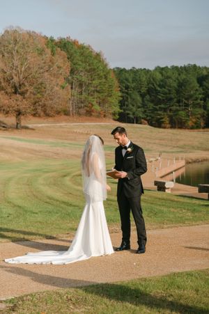 Groom in black tuxedo reads heartfelt vows to bride in white mermaid gown during outdoor first look.