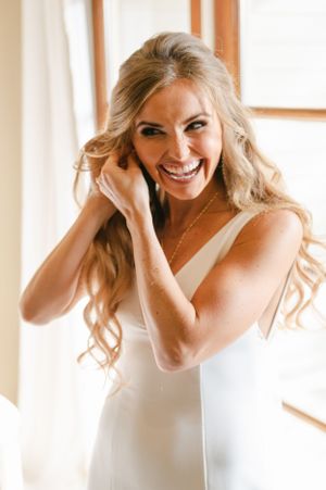 Joyful blonde bride in sleek white gown puts on earrings while smiling brightly by a sunny window.