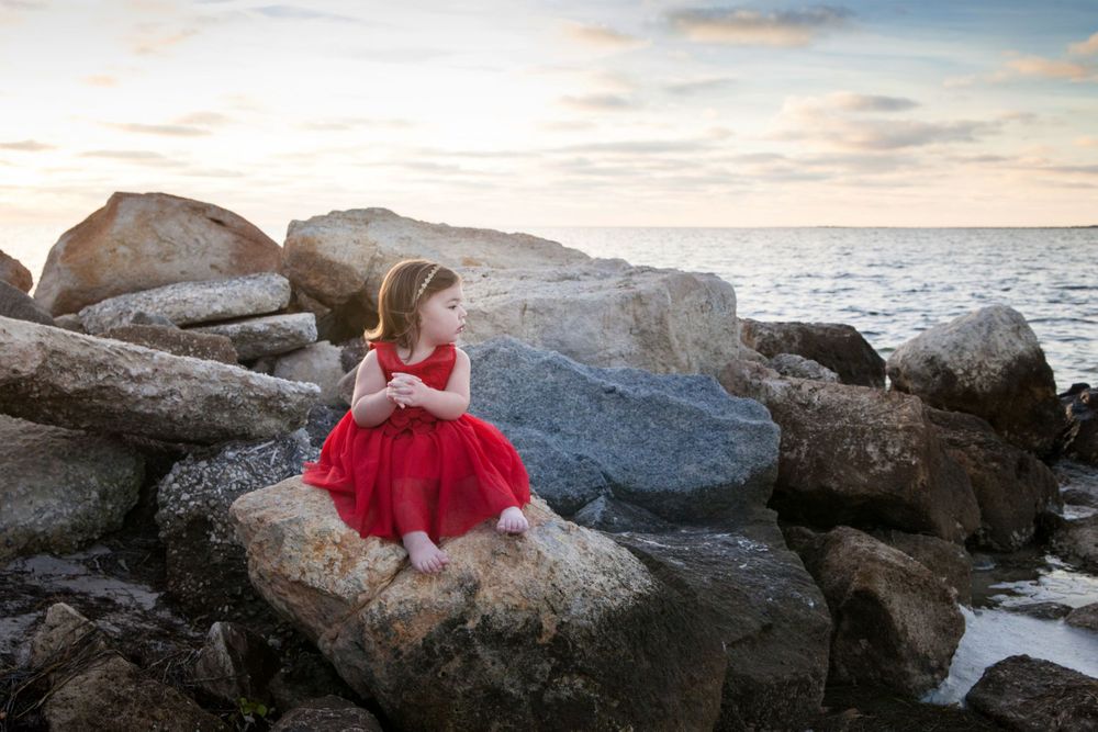 Little girl in red dress sitting on a big rock at the waters edge at sunset.